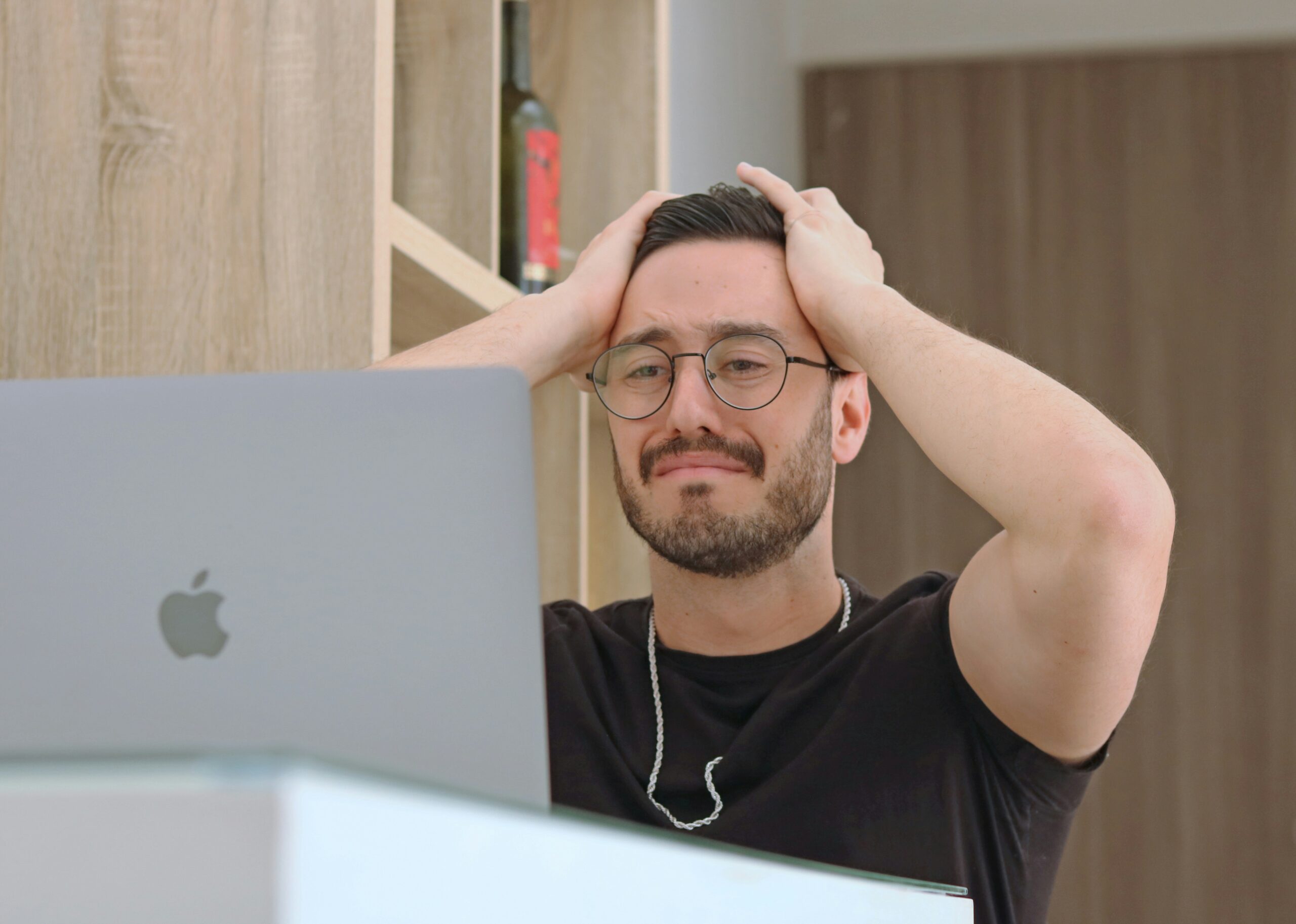 A man looking into his macbook screen with a distraught expression on his face and both of his hands pressed against his head